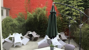 Un patio al aire libre con mesas y sillas blancas, un paraguas verde y un muro de ladrillo rojo al fondo.