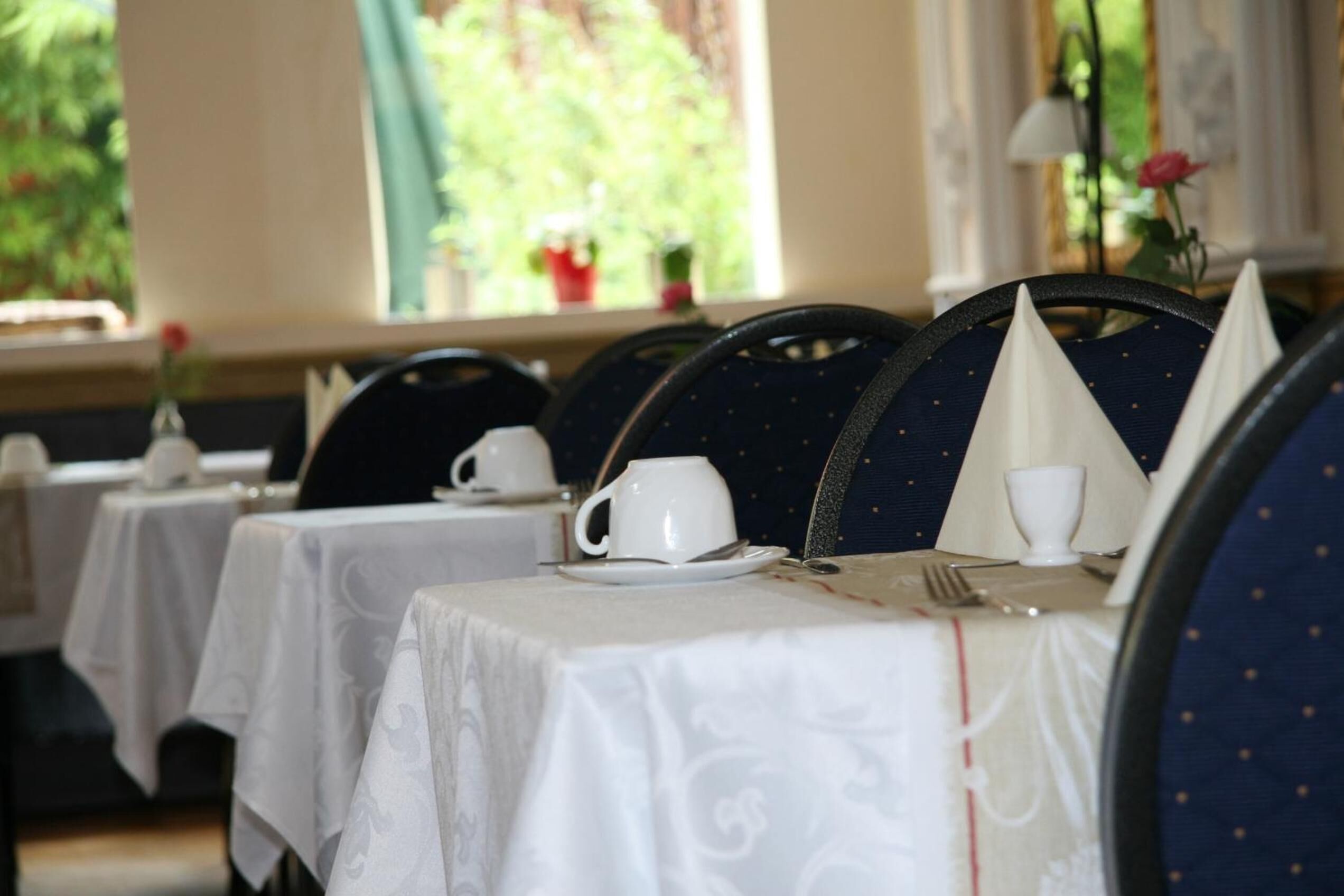 Un comedor con mesas dispuestas para un servicio de té, sillas con tela azul oscuro y lunares dorados, y una ventana que ofrece vistas a la vegetación.