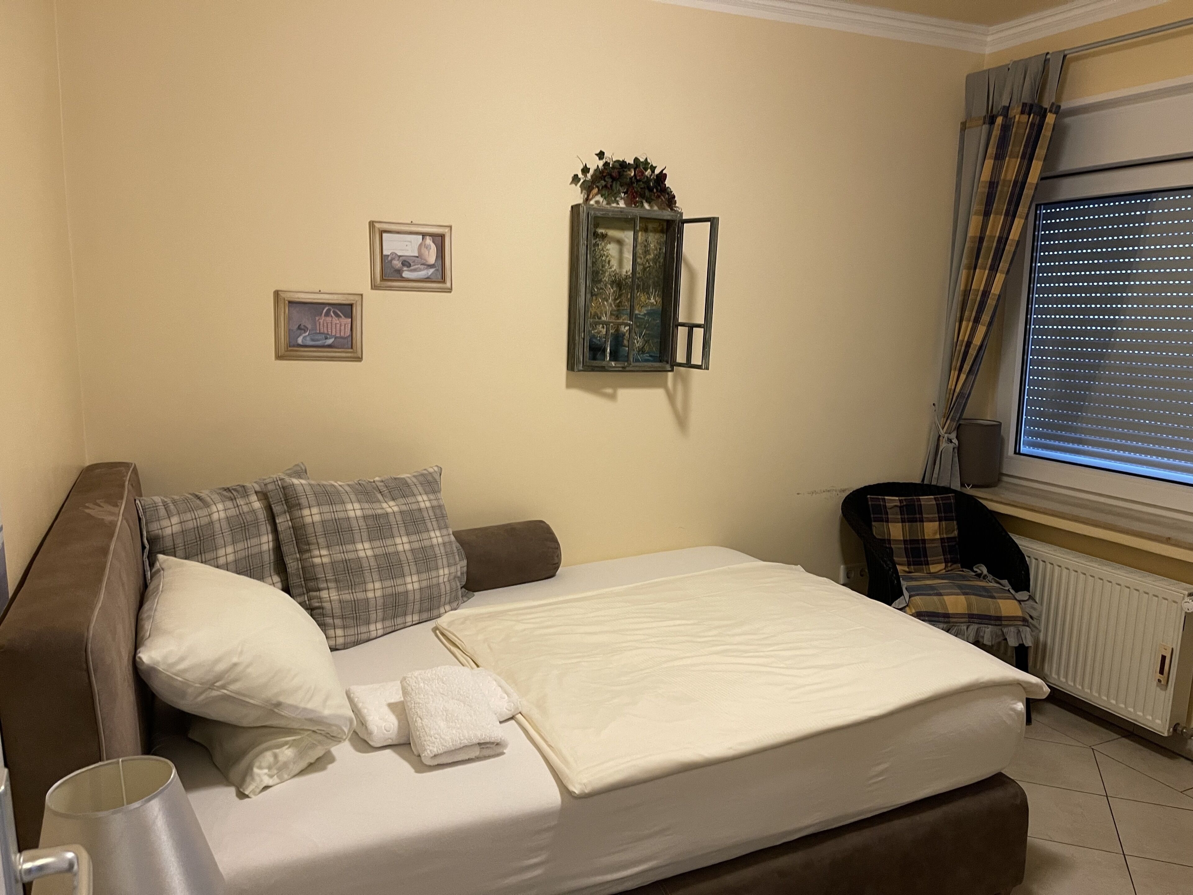 A neatly made bed with a white comforter and pillows, a small table with a lamp, and a window with blinds.