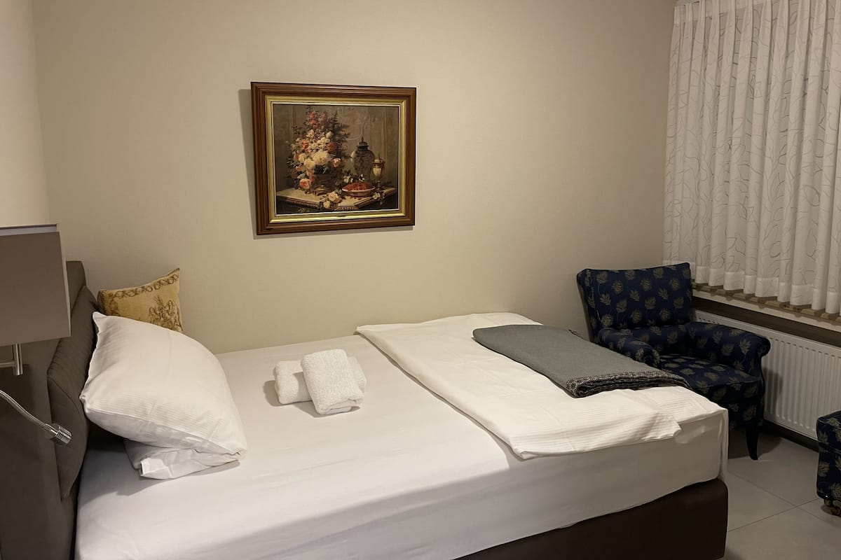 A neatly made bed with white linens, a painting of a floral arrangement on the wall, a blue patterned armchair, and a window with white curtains.