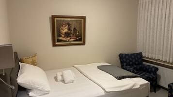 A neatly made bed with white linens, a painting of a floral arrangement on the wall, a blue patterned armchair, and a window with white curtains.