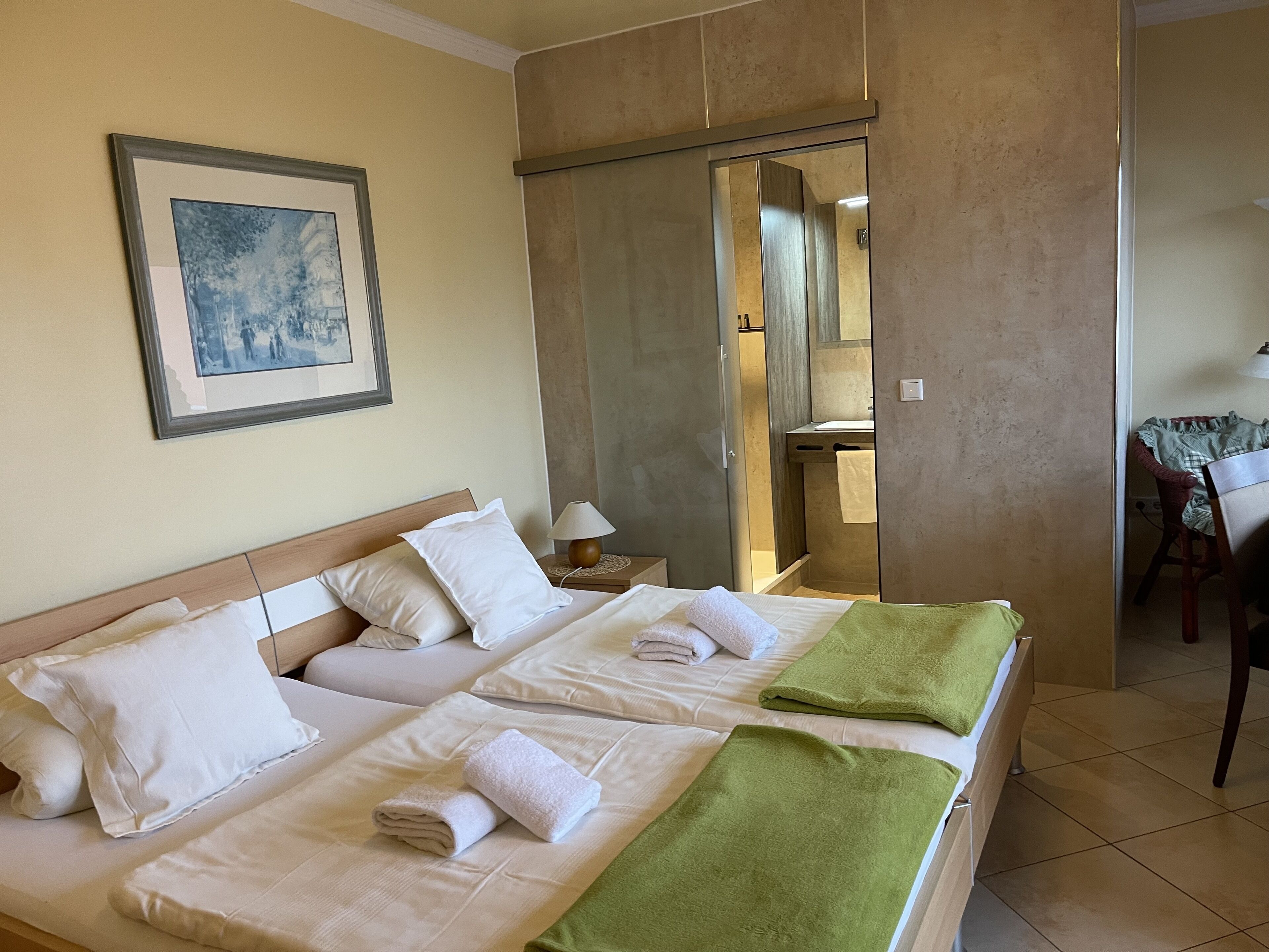A double bed with white linens and a green throw blanket, a bedside table with a lamp, and a framed picture on the wall.