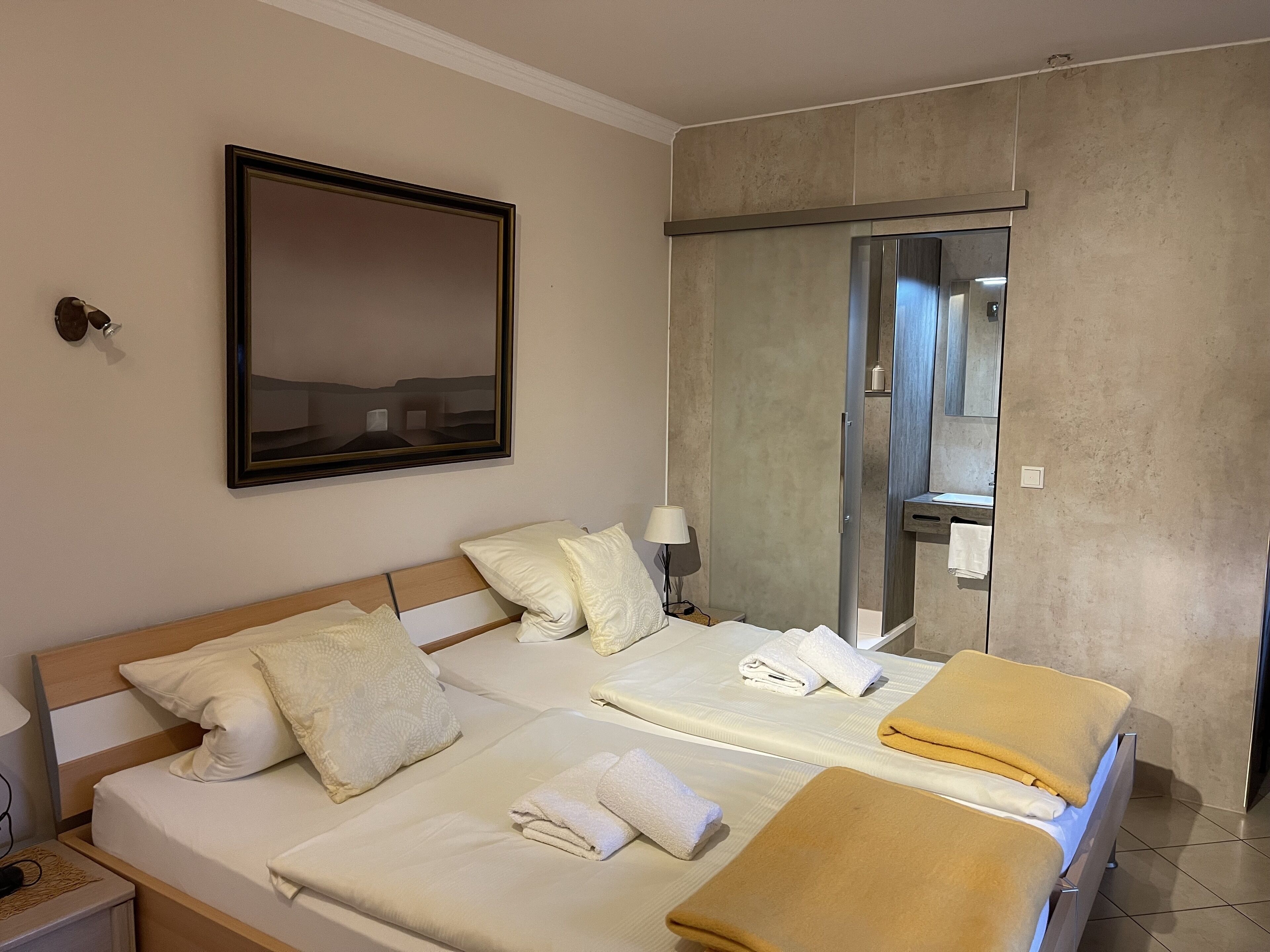 A hotel room with two beds, a large mirror, a bedside table with a lamp, and a bathroom visible through a glass door.