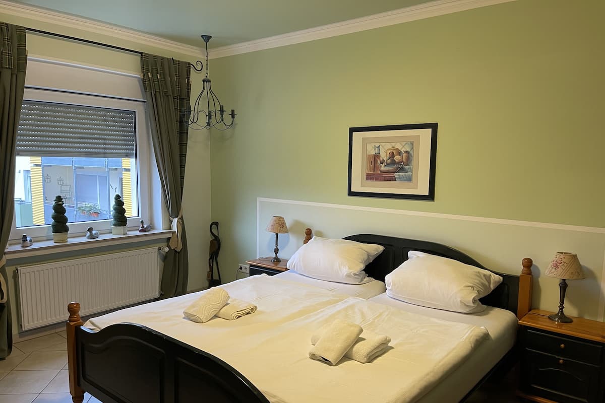 A neatly made bed with white linens and towels, a bedside table with a lamp, a framed picture on the wall, and a window with curtains.