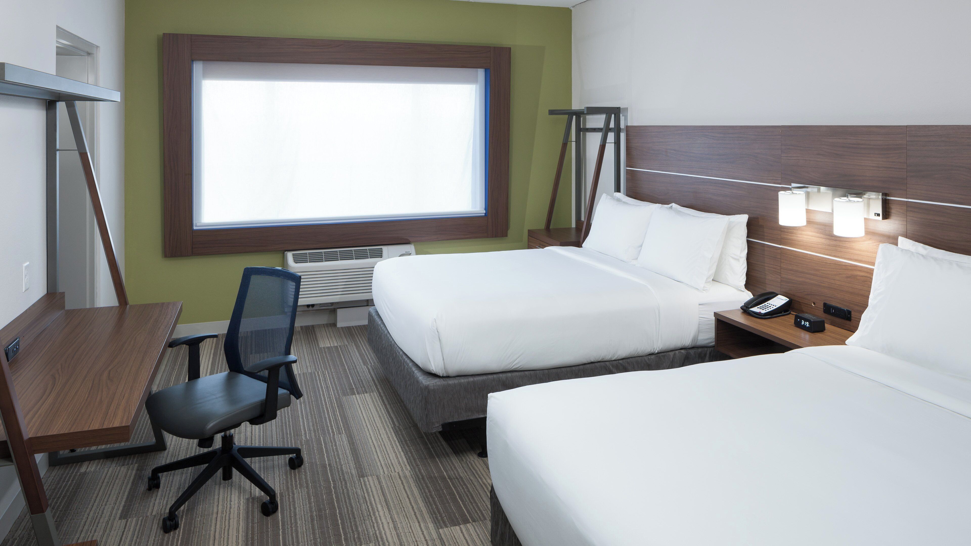 A hotel room with a desk, two beds, a chair, a window, and a nightstand with a phone and lamp.