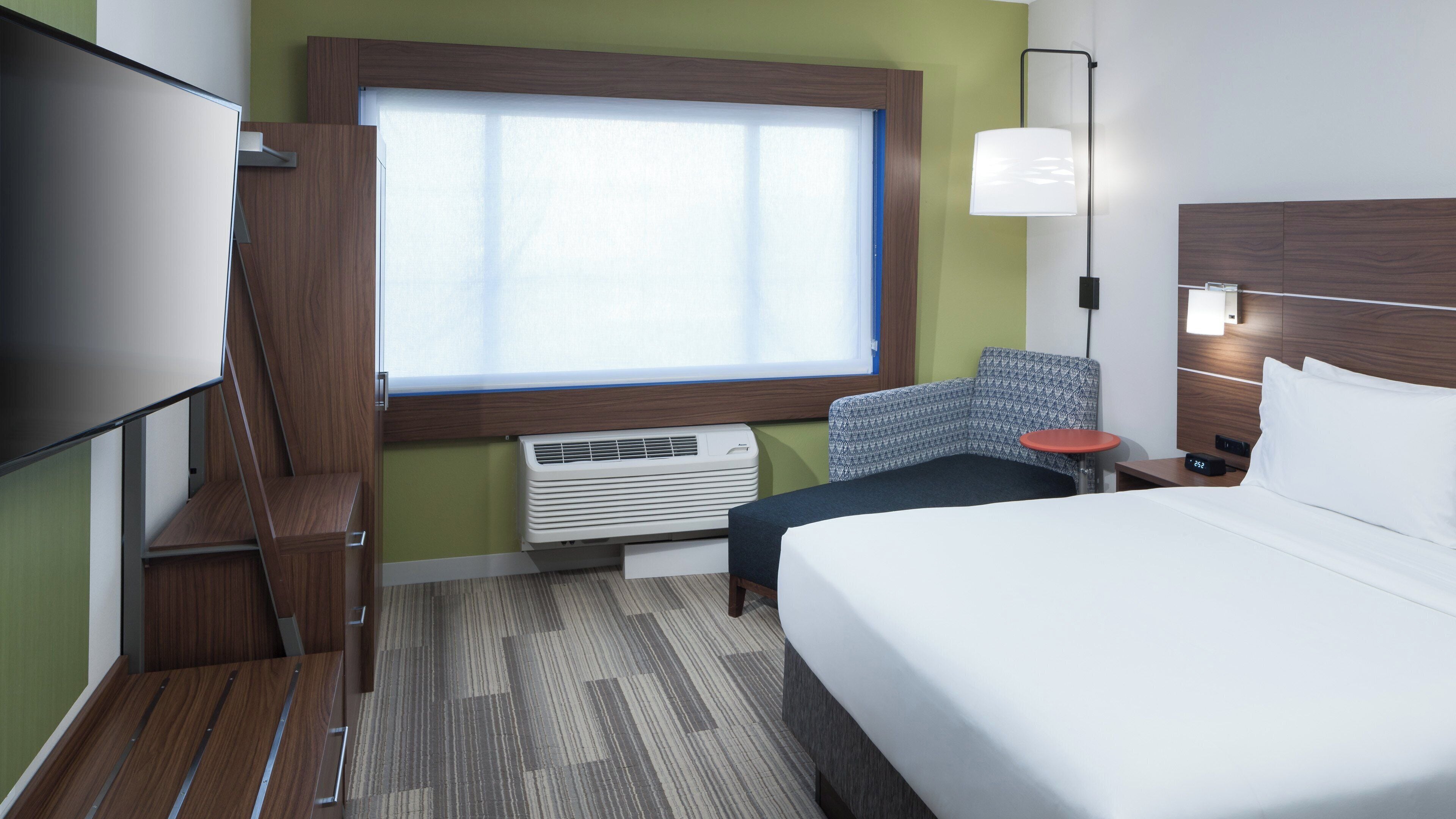 A hotel room with a bed, a chair, a TV, a window, an air conditioner, and a bedside table with a lamp.