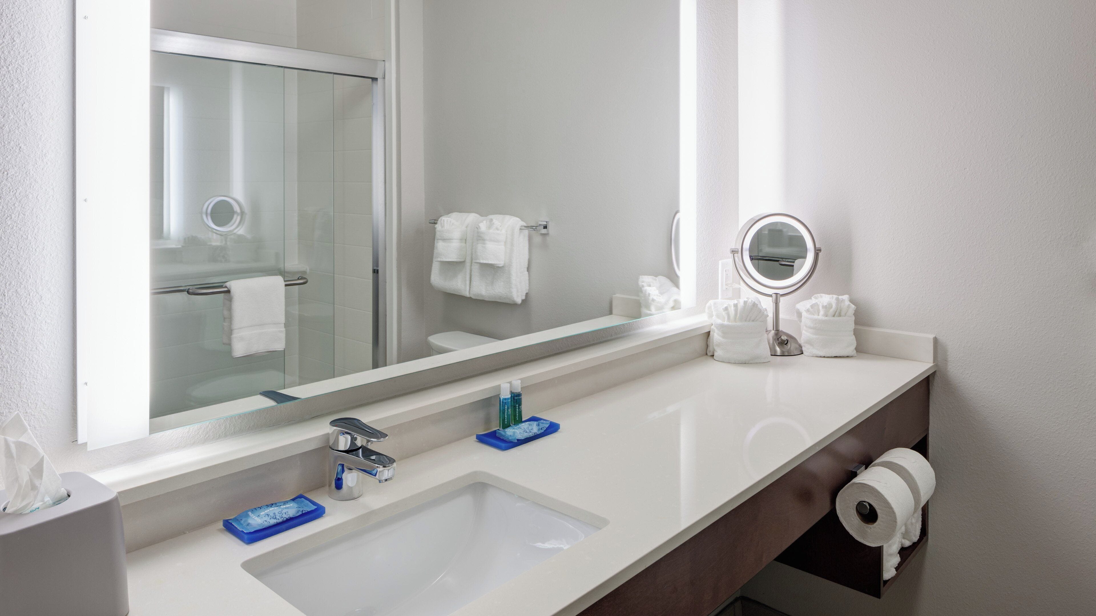 Un baño con lavamanos blanco, un espejo, toallero y dispensador de papel higiénico.