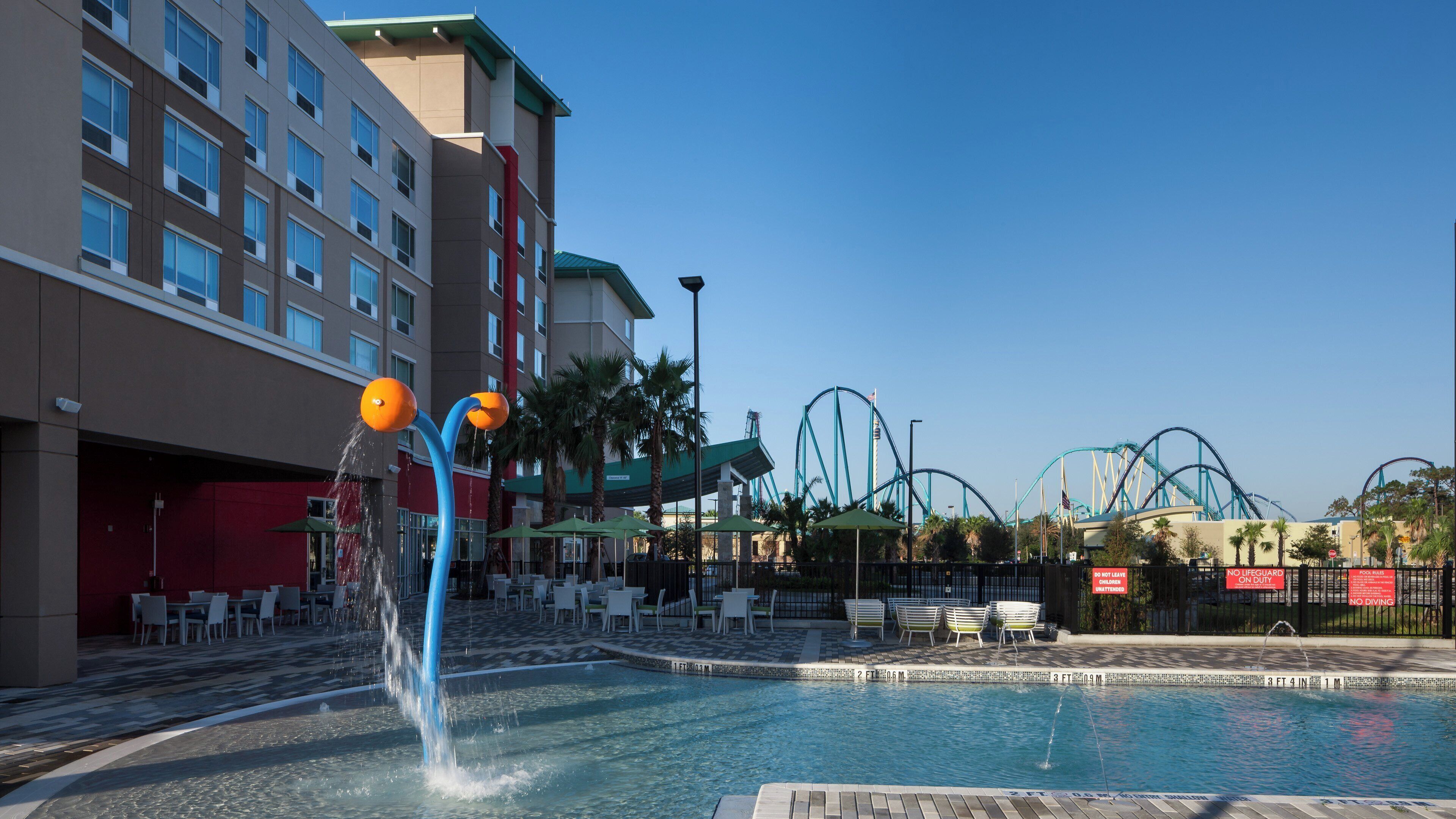 A hotel pool area with a water feature, poolside chairs, and a view of nearby amusement park rides.