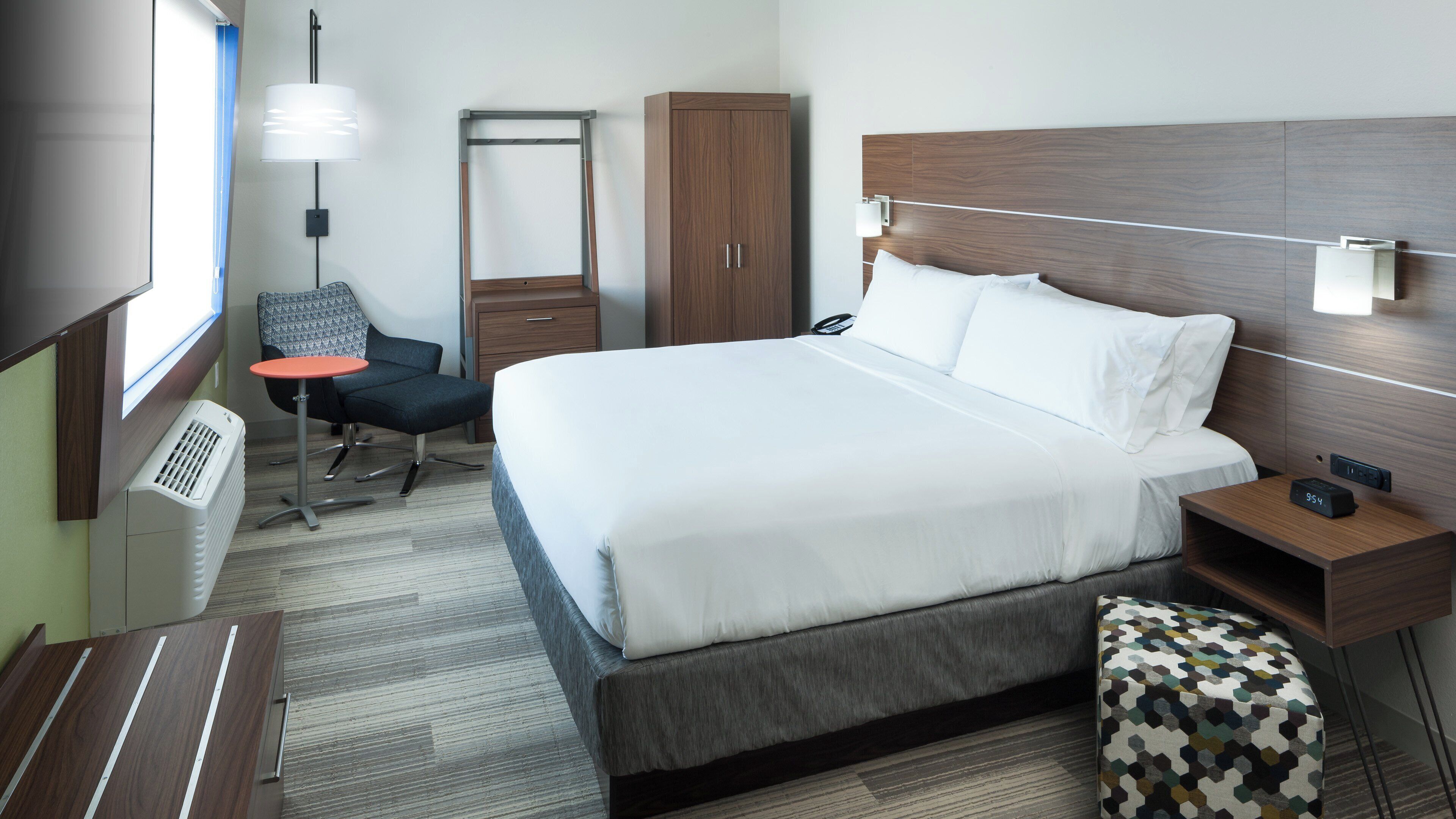 A hotel room with a bed, bedside table, chair, wardrobe, desk, lamp, and a window.