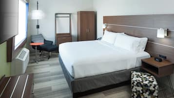 A hotel room with a bed, bedside table, chair, wardrobe, desk, lamp, and a window.