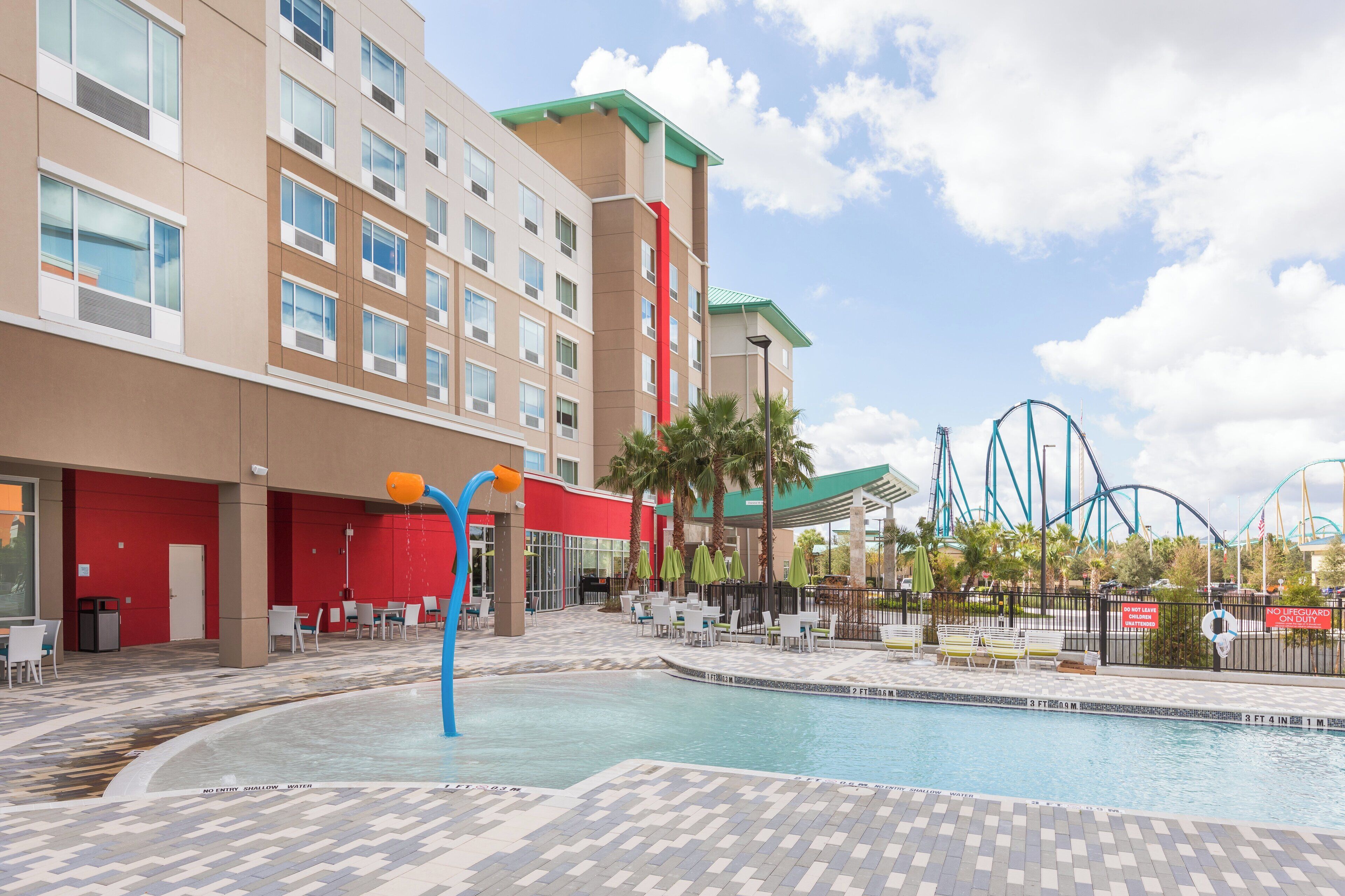 A hotel with a swimming pool, outdoor seating, and a view of a roller coaster.