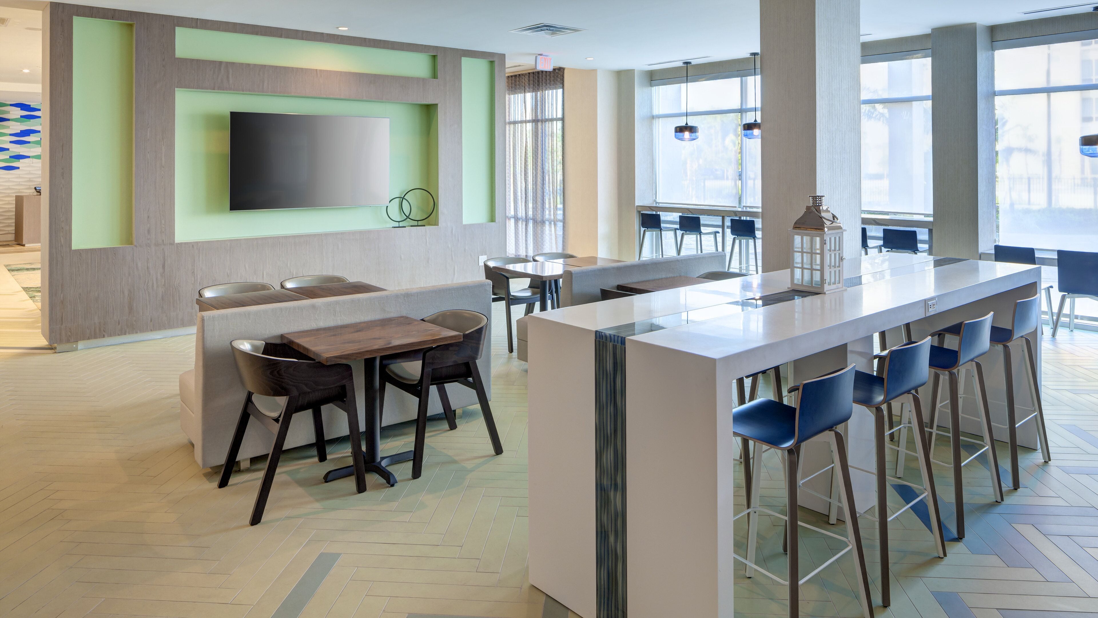 Een moderne bar met een lange toonbank en hoge krukken, een zithoek met tafels en stoelen, en een grote flatscreen aan de muur.