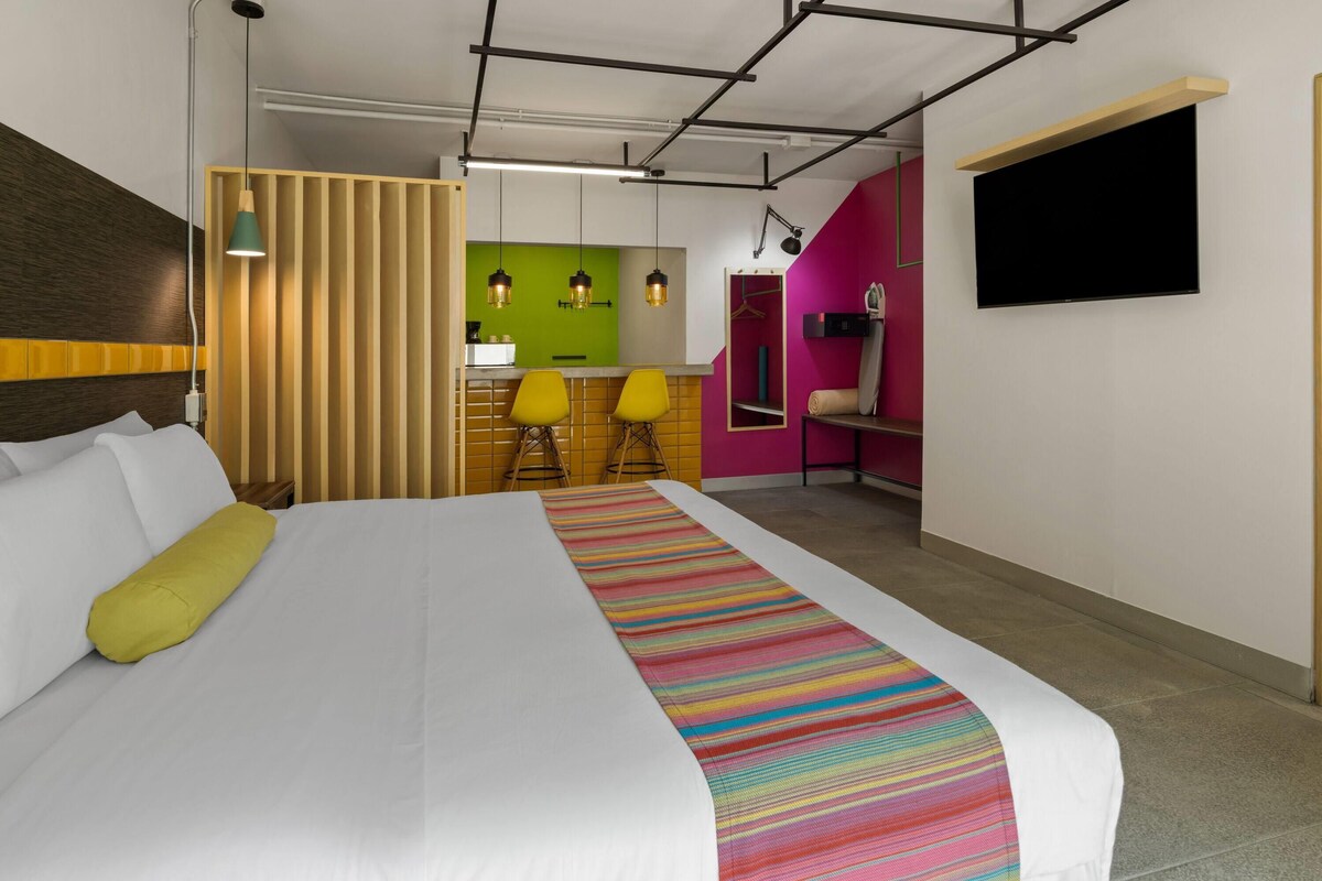 A hotel room with a large bed, a TV mounted on the wall, and a kitchenette area with a green wall and yellow chairs.