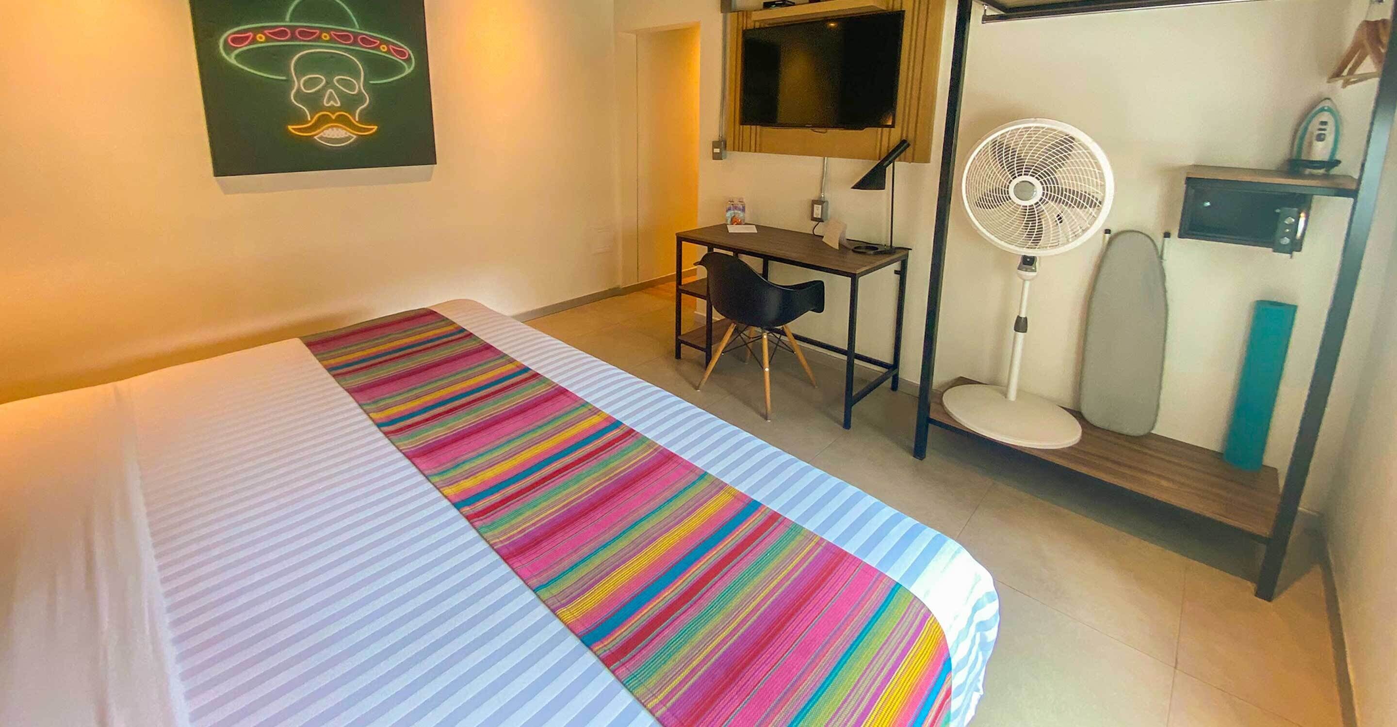 A hotel room with a bed, a desk, a chair, a desk fan, and a TV.