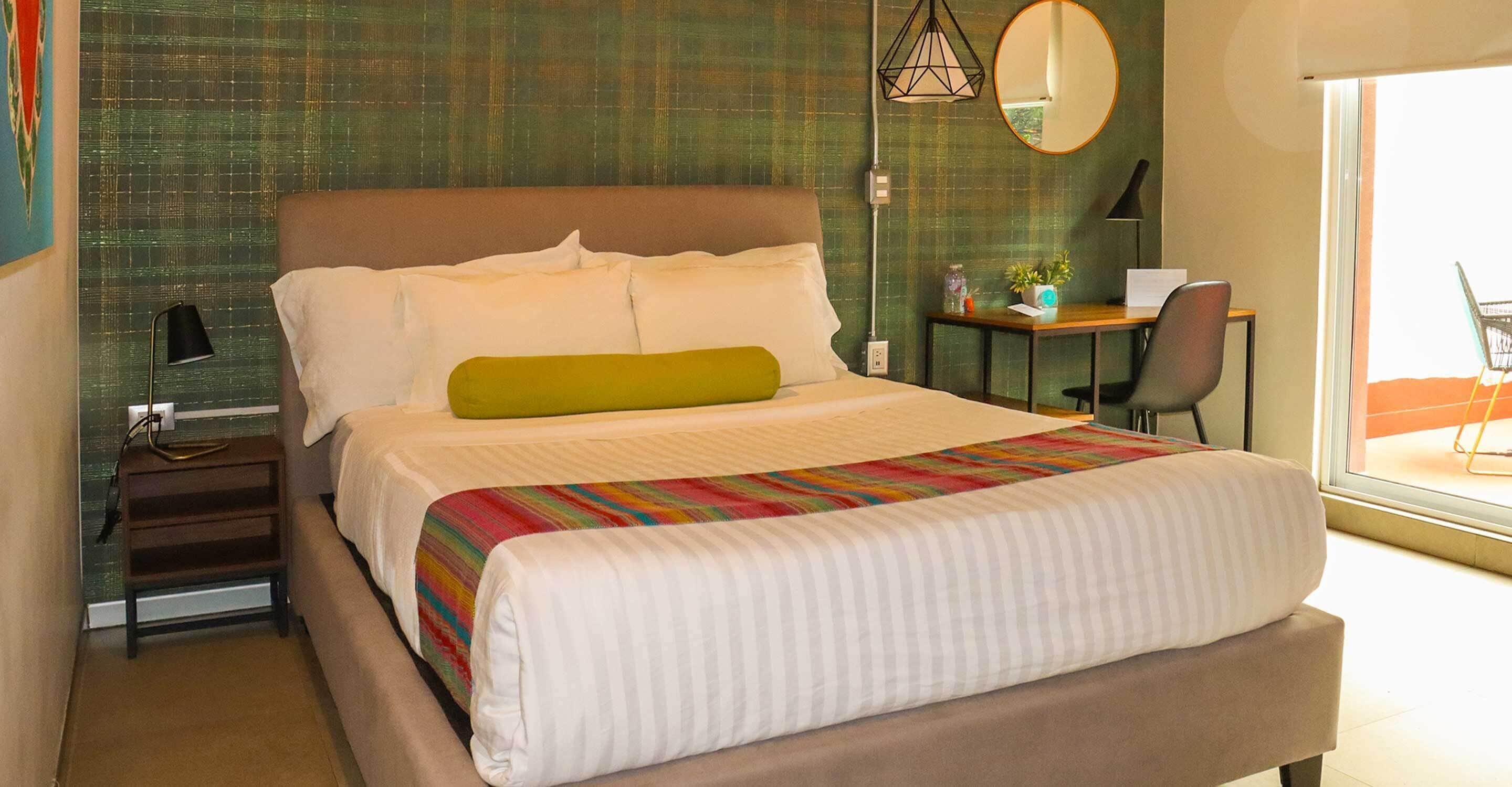 A neatly made bed with a colorful striped blanket, a green pillow, and white pillows. There is a desk with a computer and a plant, and a chair. The room has a green textured wall and a large window.