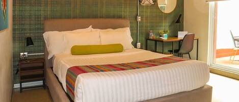 A neatly made bed with a colorful striped blanket, a green pillow, and white pillows. There is a desk with a computer and a plant, and a chair. The room has a green textured wall and a large window.