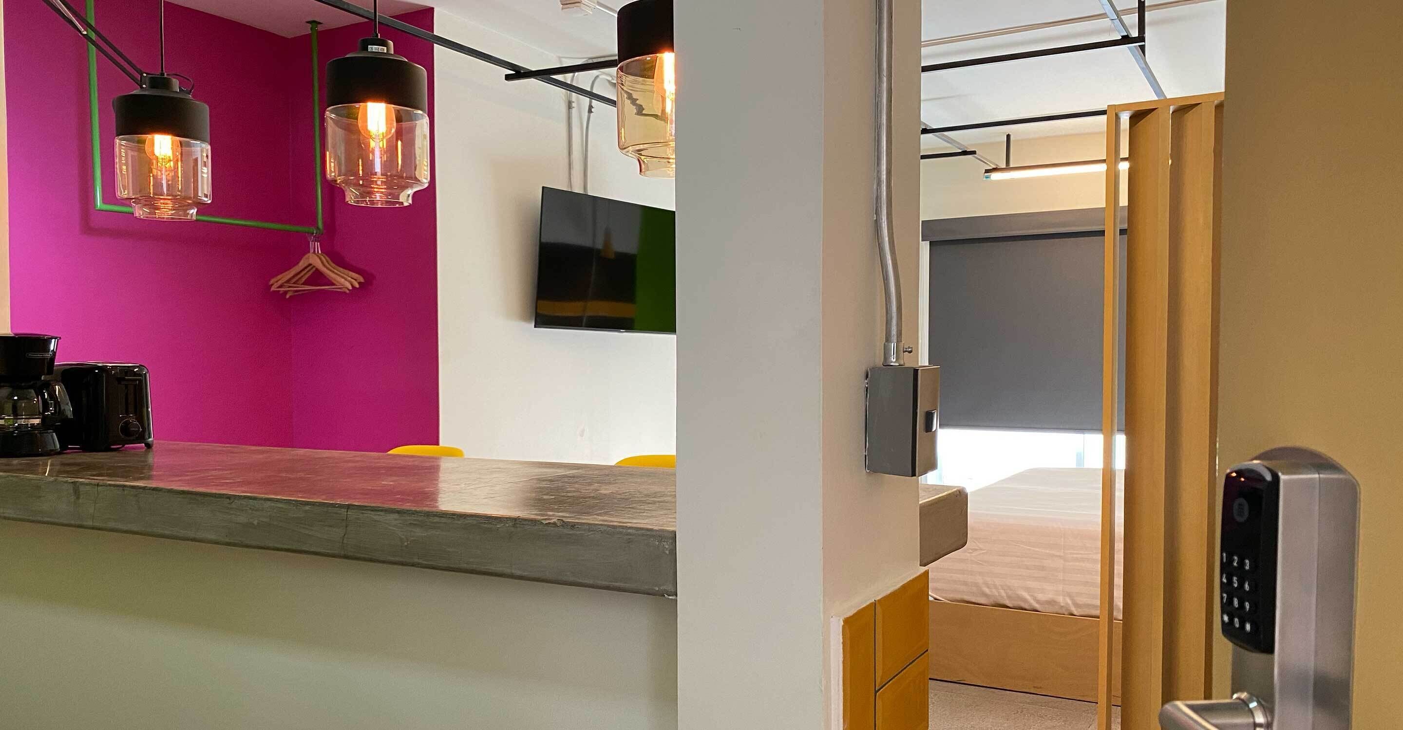 A modern hotel room with a bed, a desk with a coffee machine, and a bright pink wall.