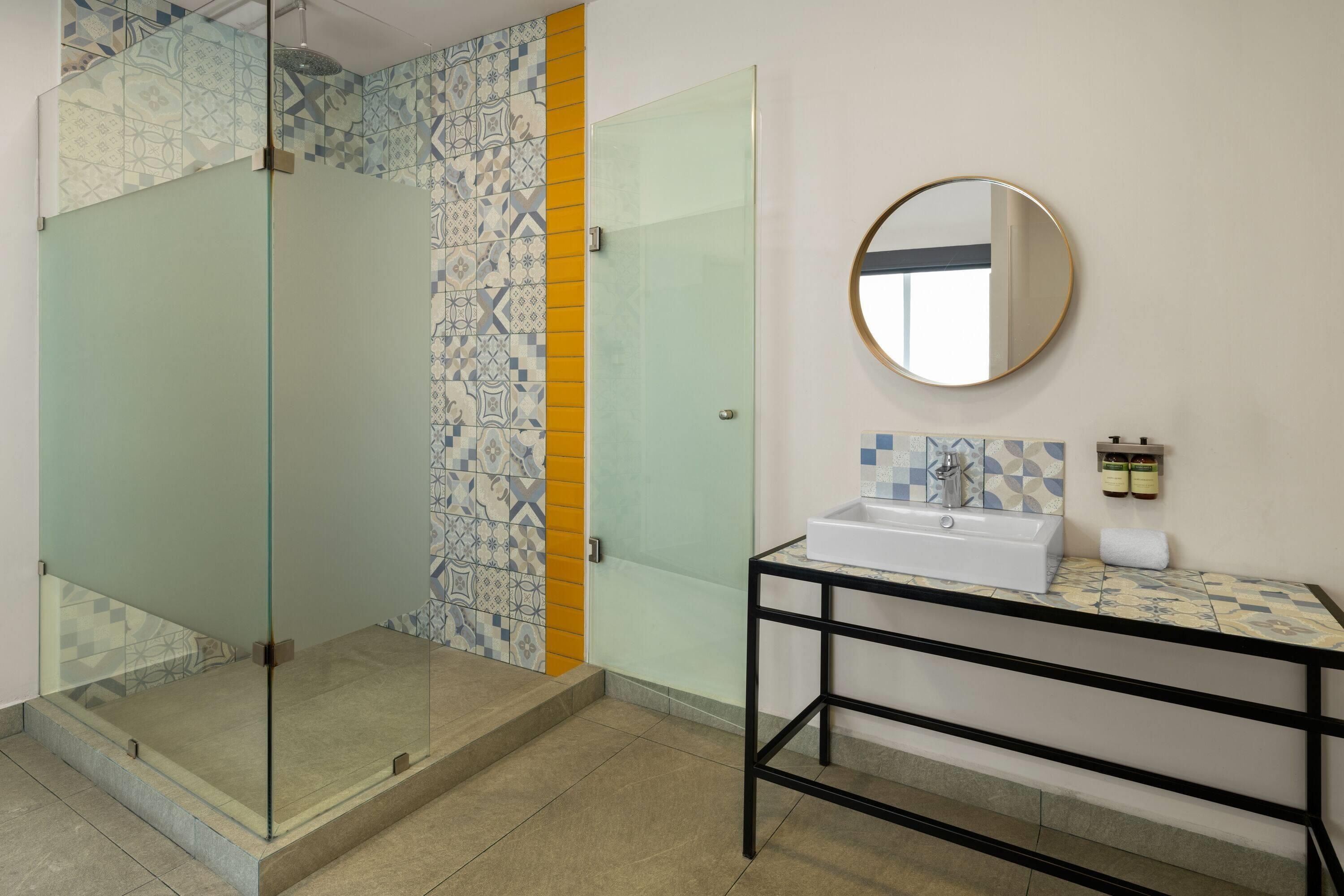 Un baño con cabina de ducha de vidrio, pared revestida con un diseño geométrico, lavabo blanco con espejo redondo y una pequeña mesa con artículos de aseo.
