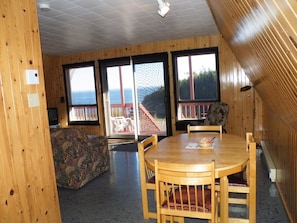 Ein Holzinterieur mit einem Esstisch, Stühlen, einer gemusterten Couch und großen Fenstern mit Blick ins Freie.