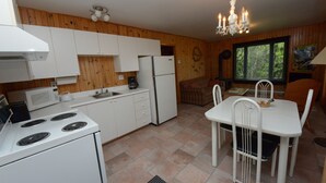 A compact kitchen with white appliances, a dining area with a white table and chairs, and a chandelier. - Chalets de l'anse a Jos et anse a Yves (Essipit)