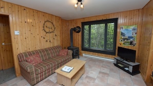 A cozy room with wooden walls, a plaid sofa, a small coffee table, a fireplace, and a large window. - Chalets de l'anse a Jos et anse a Yves (Essipit)