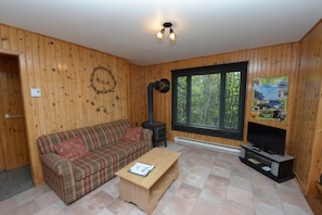 Ein gemütliches Zimmer mit Holzwänden, einem Karomuster-Sofa, einem kleinen Couchtisch, einem Kamin und einem großen Fenster.