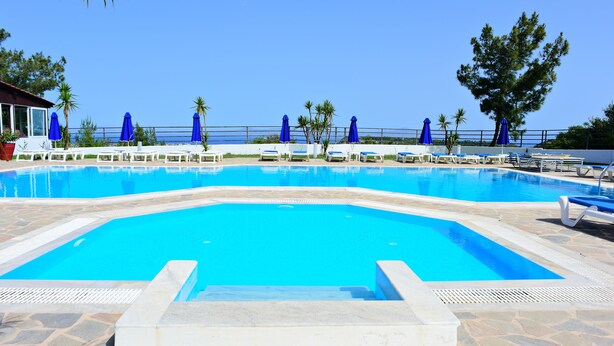 Une piscine entourée de dalles de pierre et de palmiers, avec des chaises longues blanches et des parasols bleus.