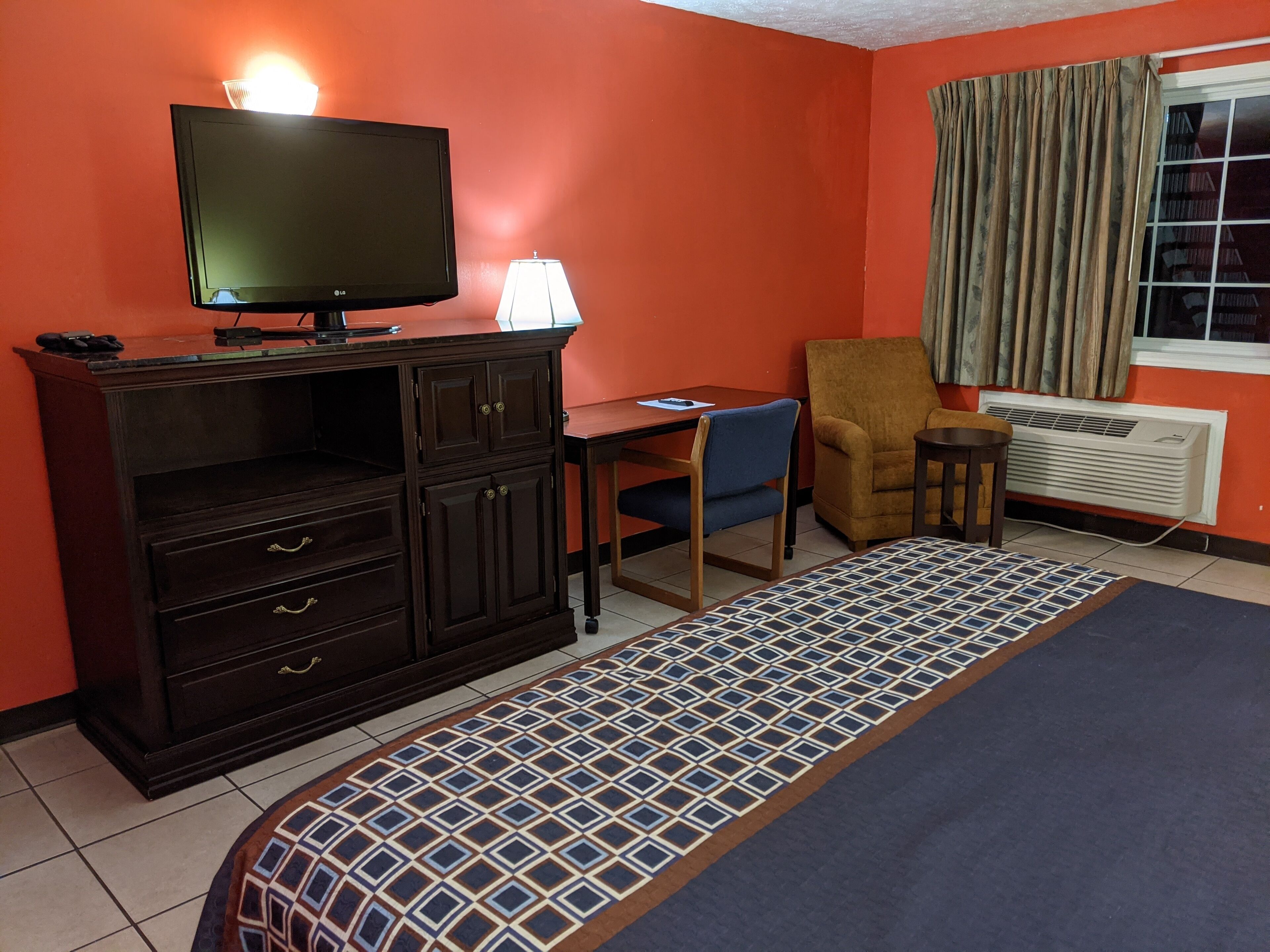 A hotel room with a bed, a desk, a chair, a TV, a lamp, and a window with curtains.