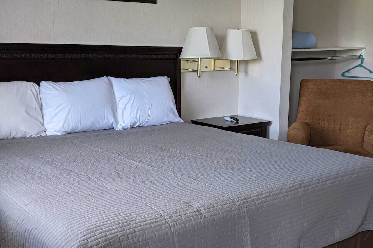 A neatly made bed with white pillows and a grey quilt, a bedside table with a lamp, and a brown chair.