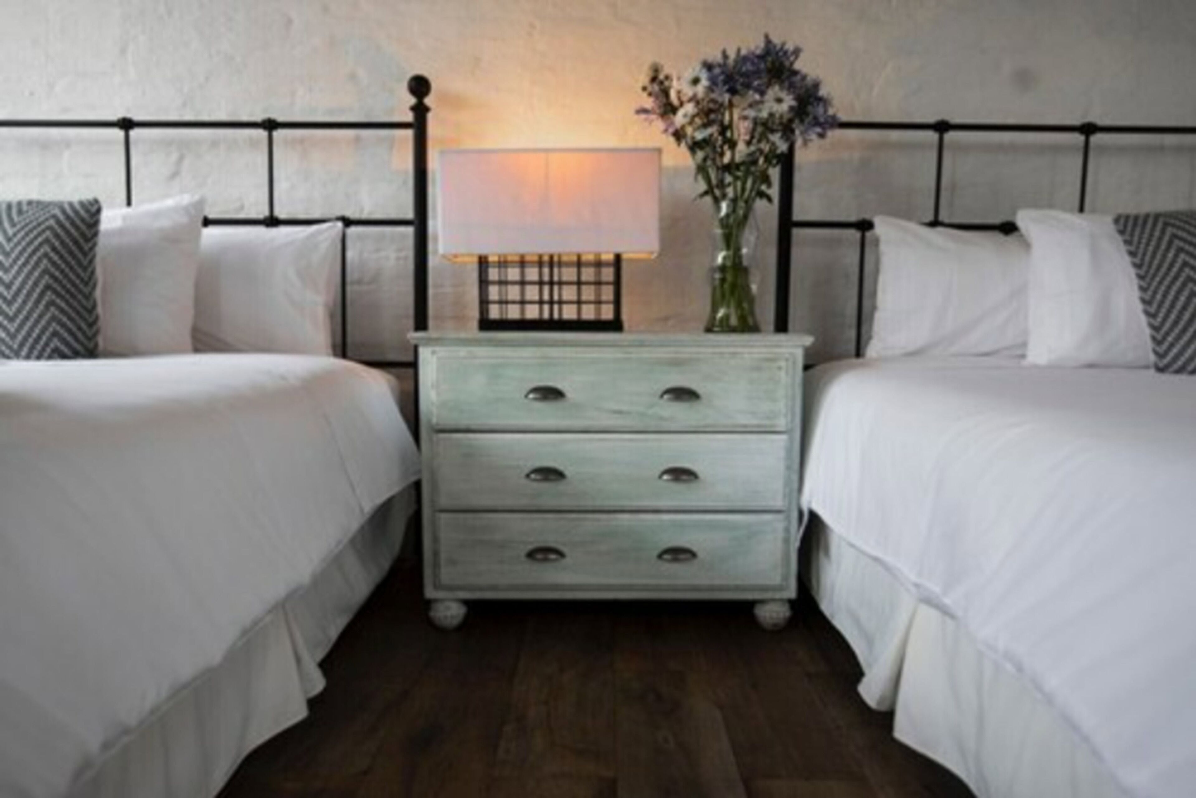 A bedroom with two beds, a nightstand with a lamp, and a vase with flowers.