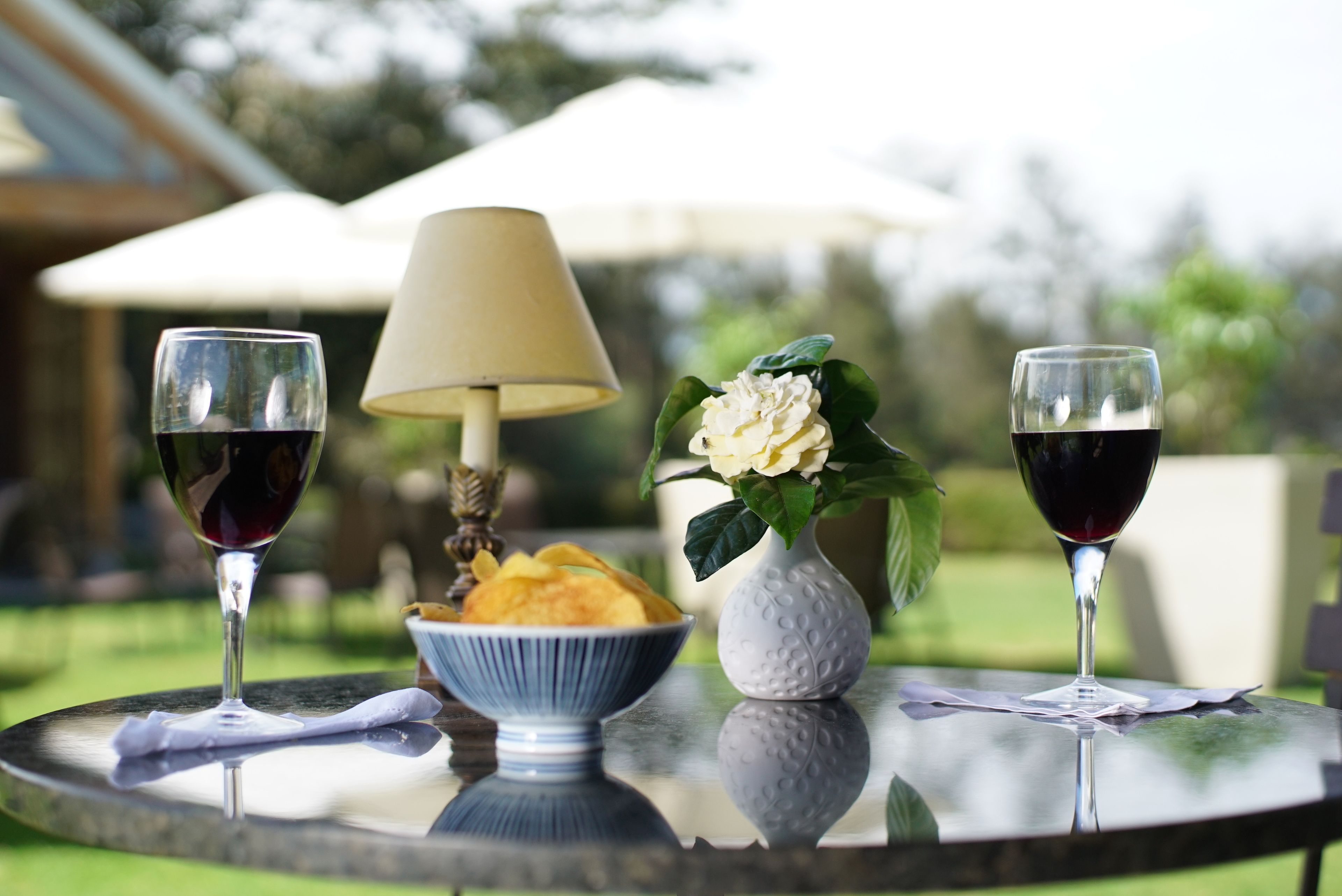 A round table with two wine glasses, a bowl of snacks, a lamp, and a vase with flowers.