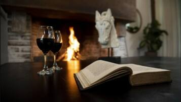 Une cheminée avec un feu allumé, deux verres de vin rouge et un livre ouvert sur une table.
