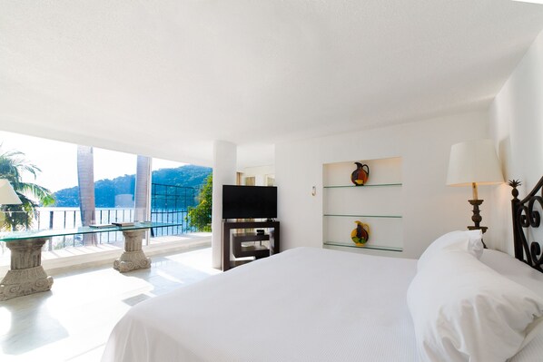 A hotel room with a large bed, a television, a glass table, and a balcony with a view of the mountains. - Exclusiva Villa Aldila frente al mar en Acapulco (Acapulco)