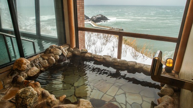 A stone hot tub with a view of the ocean through large windows. - Hotel Tappi (Sotogahama)