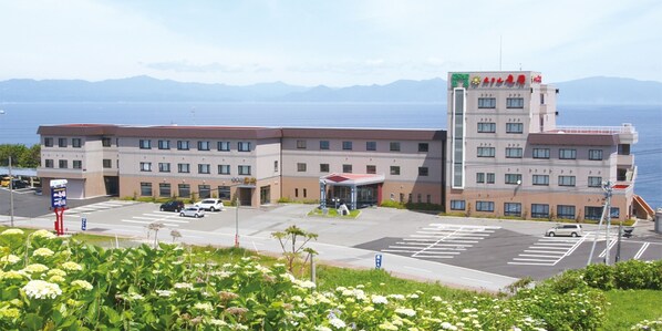 A hotel with a parking lot, a building with multiple floors, and a view of the sea in the background. - Hotel Tappi (Sotogahama)