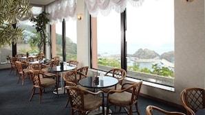 A dining area with wicker chairs and round tables, large windows with white curtains, and a view of a coastal landscape. - Hotel Tappi (Sotogahama)