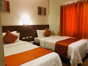 Une chambre d’hôtel avec deux lits, chacun agrémenté de linge de lit blanc et de touches de couleur orange, d’une tête de lit en bois et d’une grande fenêtre avec des rideaux.
