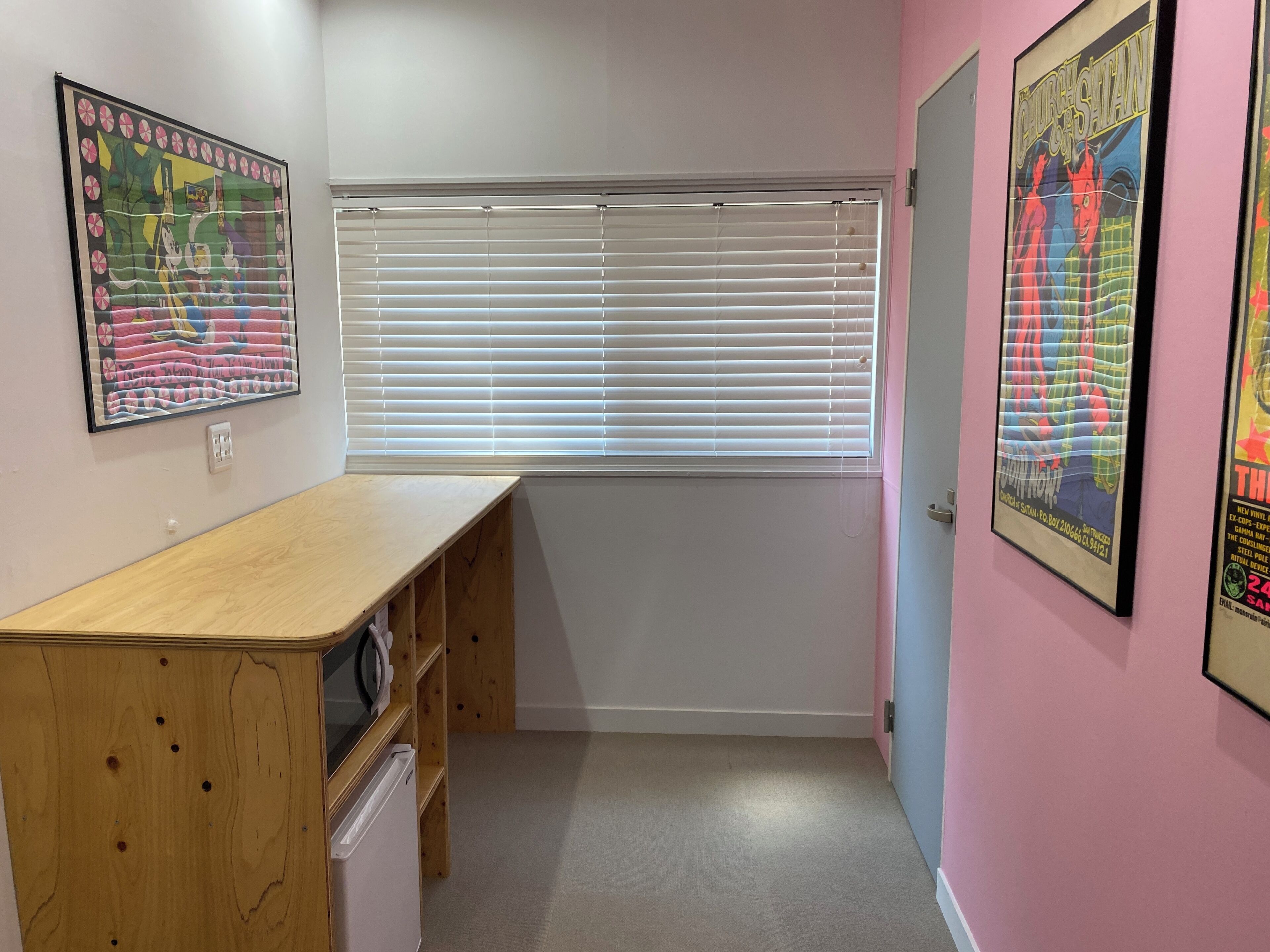 A room with a wooden desk, a window with blinds, and two framed posters on the wall.