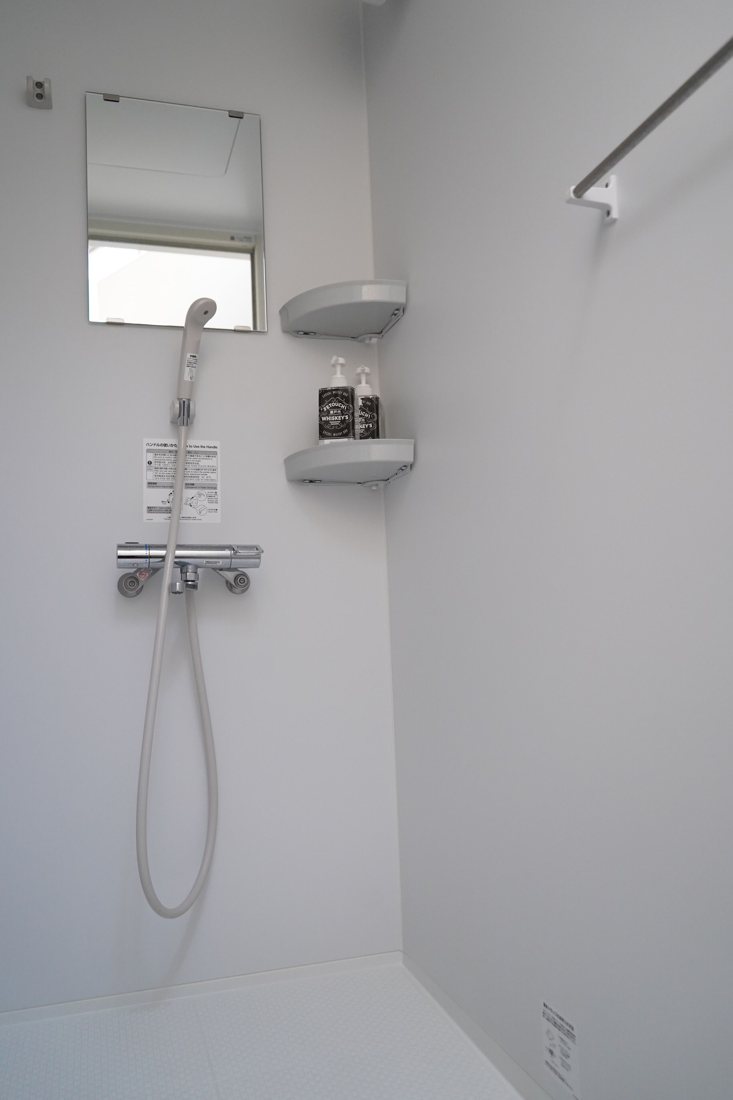 Una ducha moderna con cabezal de ducha manual y panel de control en la pared. Hay un pequeño estante con artículos de tocador y un espejo sobre la ducha.