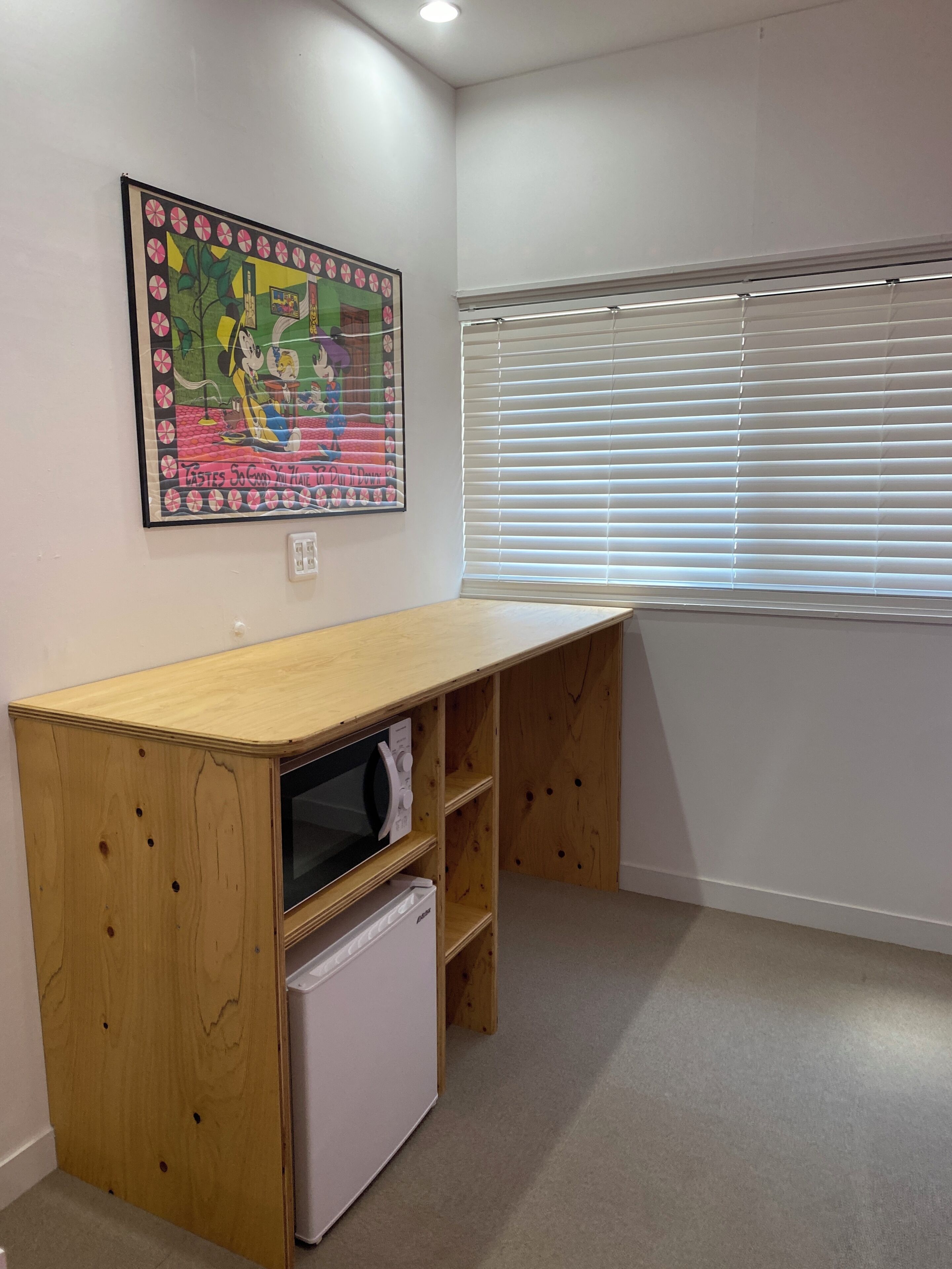 Een kamer met een houten bureau, een kleine witte koelkast, een raam met zonwering en een kleurrijk poster op de muur.