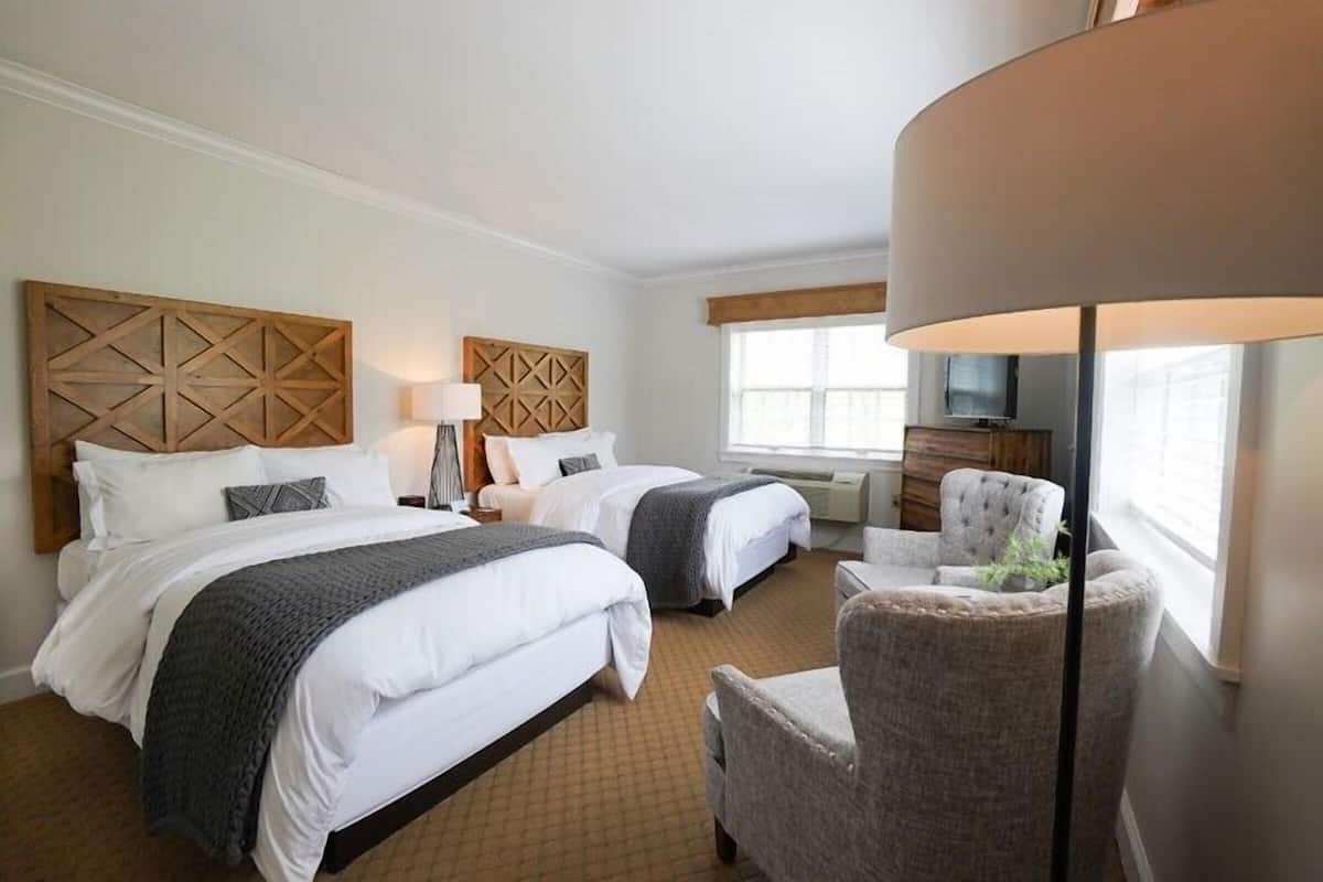 Quarto de hotel com duas camas, uma escrivaninha, uma cadeira, um abajur e uma televisão.