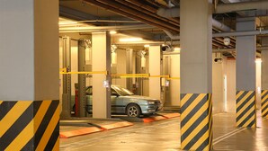 A parking garage with a car in a lift, yellow and black striped barriers, and visible structural beams and pipes. - Go Hotels North Edsa (Quezon City)
