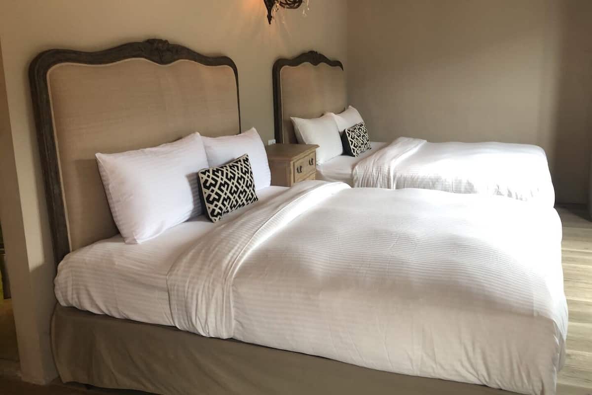 Una cama doble con sábanas blancas y almohadas estampadas, una mesita de noche y un florero con flores.