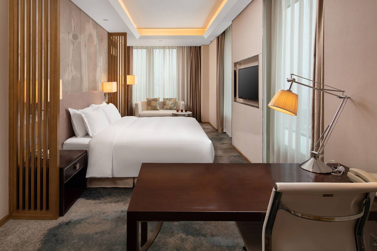 Quarto de hotel com cama, mesas de cabeceira, escrivaninha, cadeira, televisão e luminária.