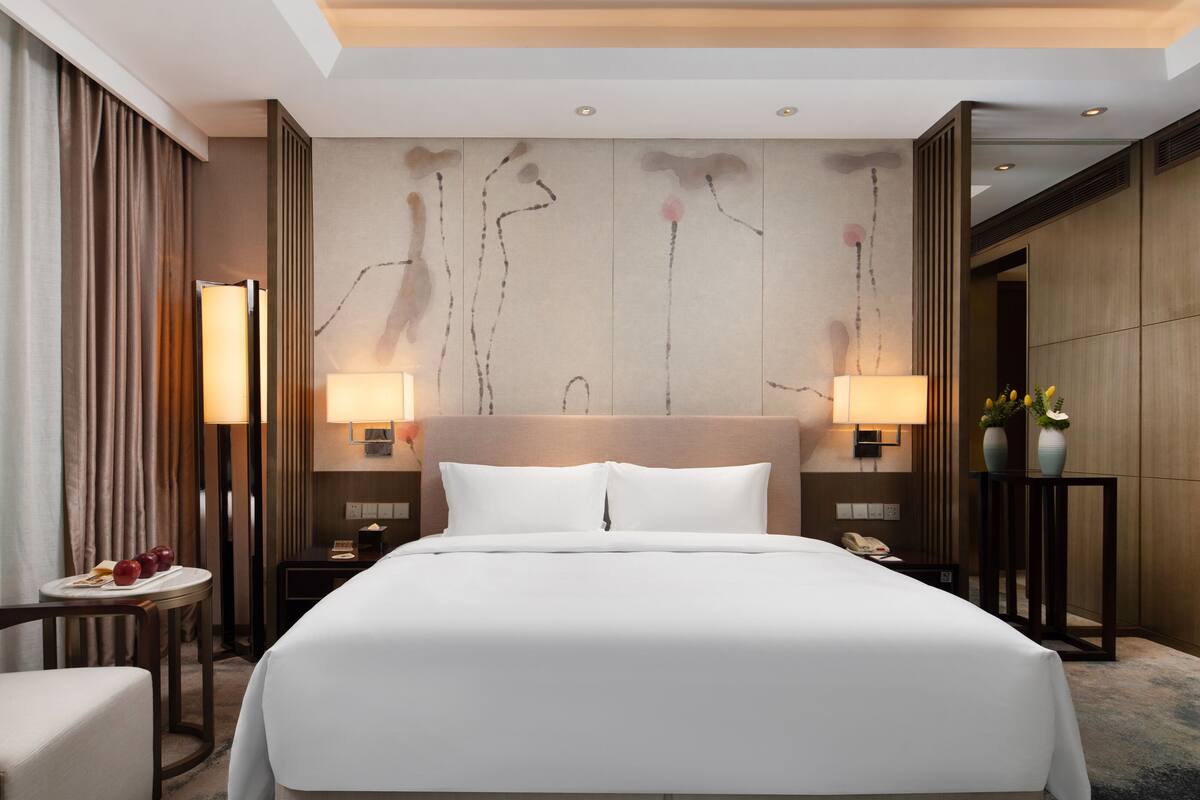 Quarto de hotel com uma cama grande, duas luminárias de cabeceira, uma mesinha de cabeceira com telefone, uma pequena mesa com um vaso de flores e uma cadeira.
