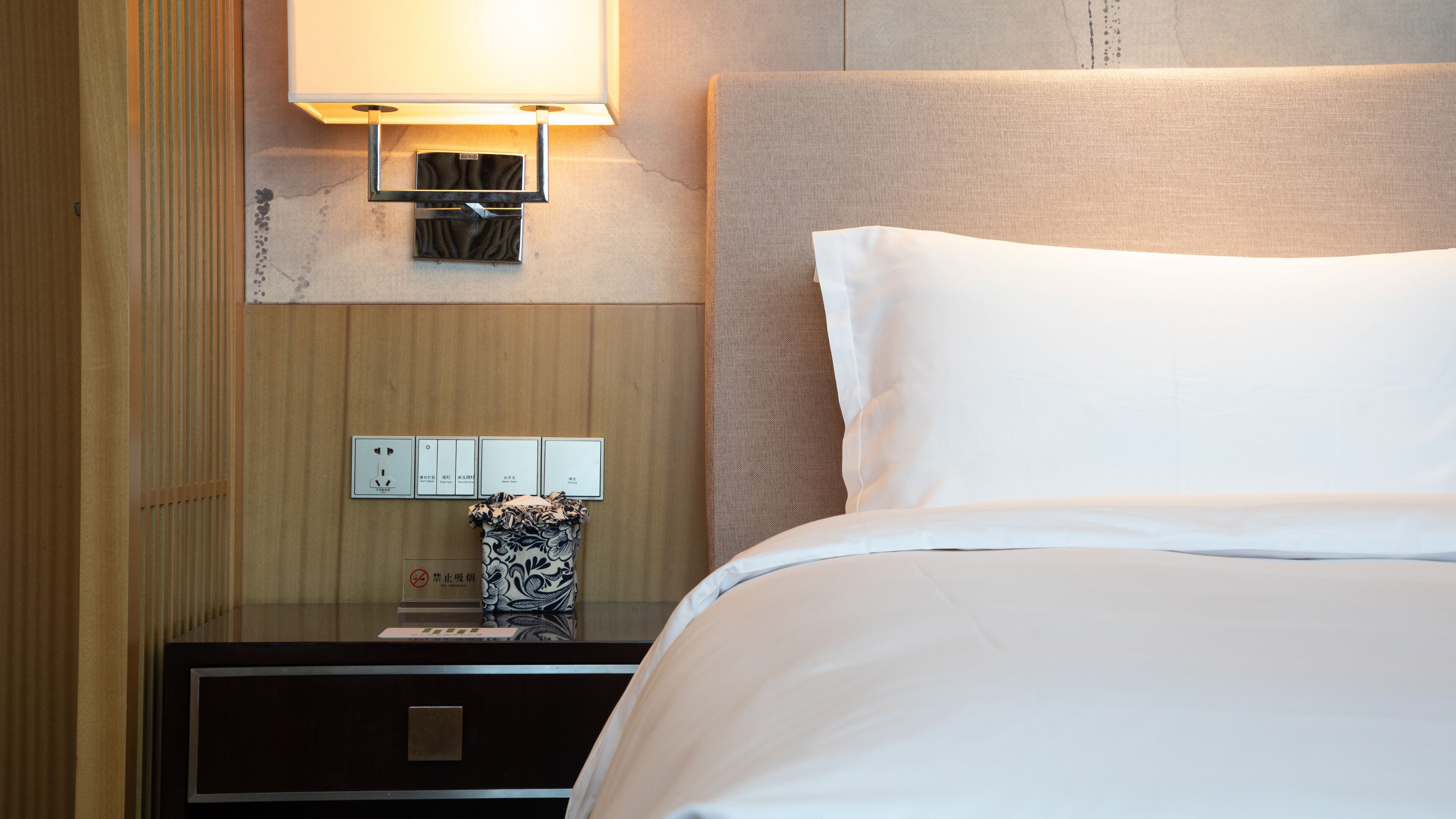 A hotel room with a bed, a nightstand, a lamp, and a wall-mounted power outlet.