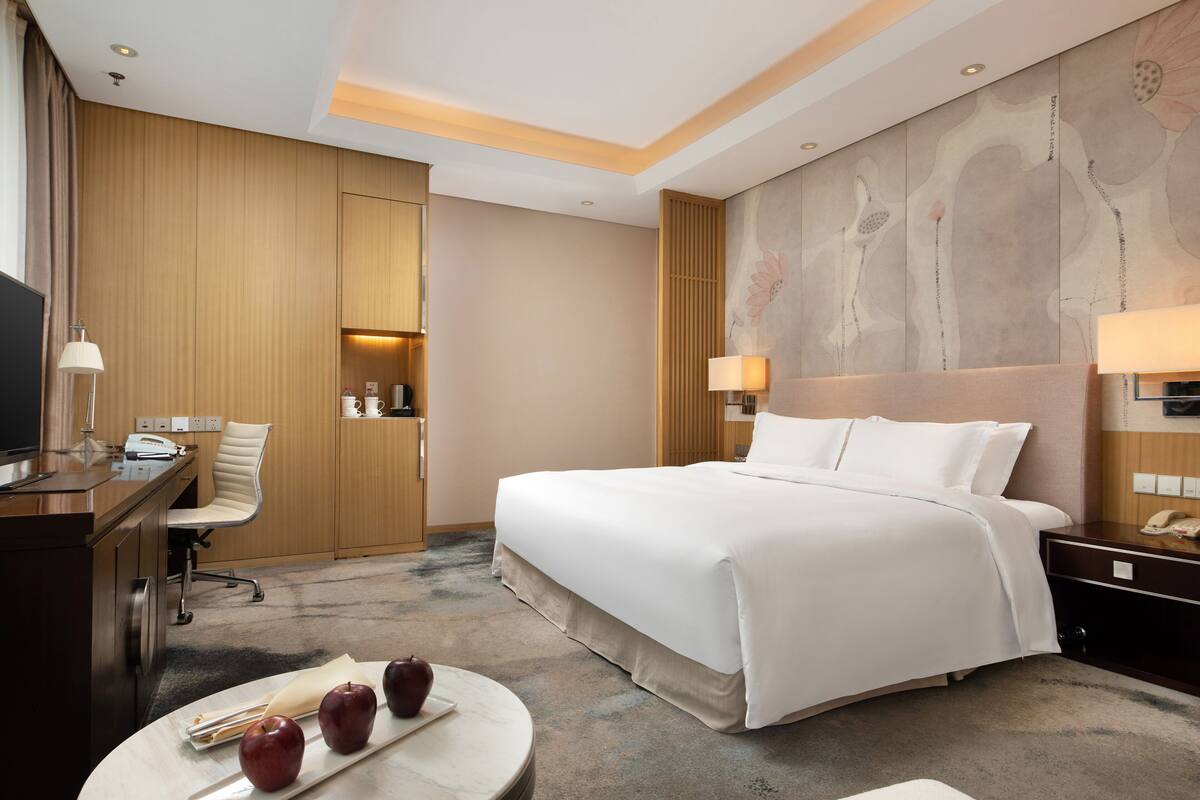 Quarto de hotel com uma cama grande, uma escrivaninha com uma lâmpada, uma cadeira e uma pequena mesa com maçãs.