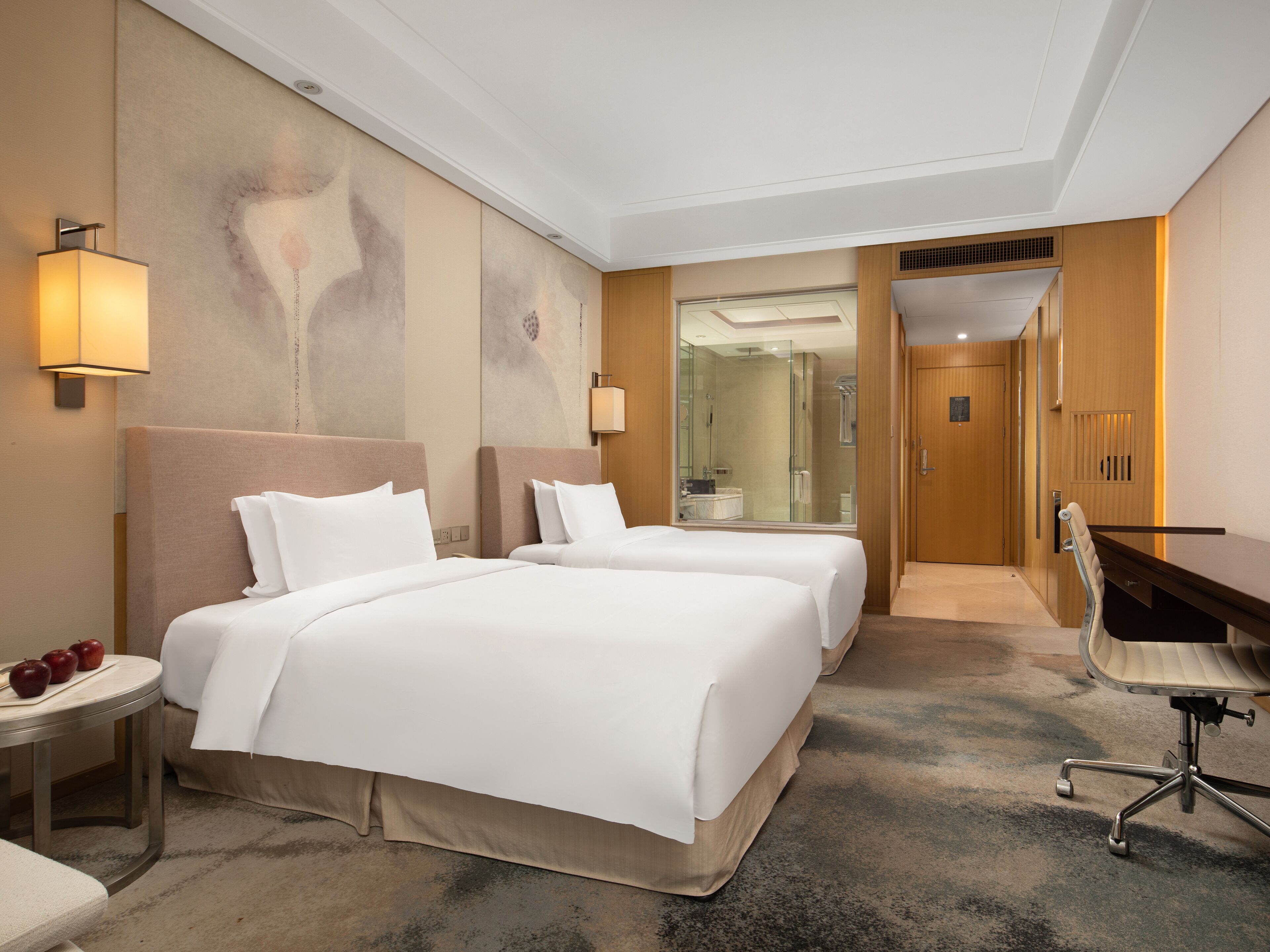 A hotel room with two beds, a desk, a chair, a small table with apples, and a bathroom visible through a glass door.