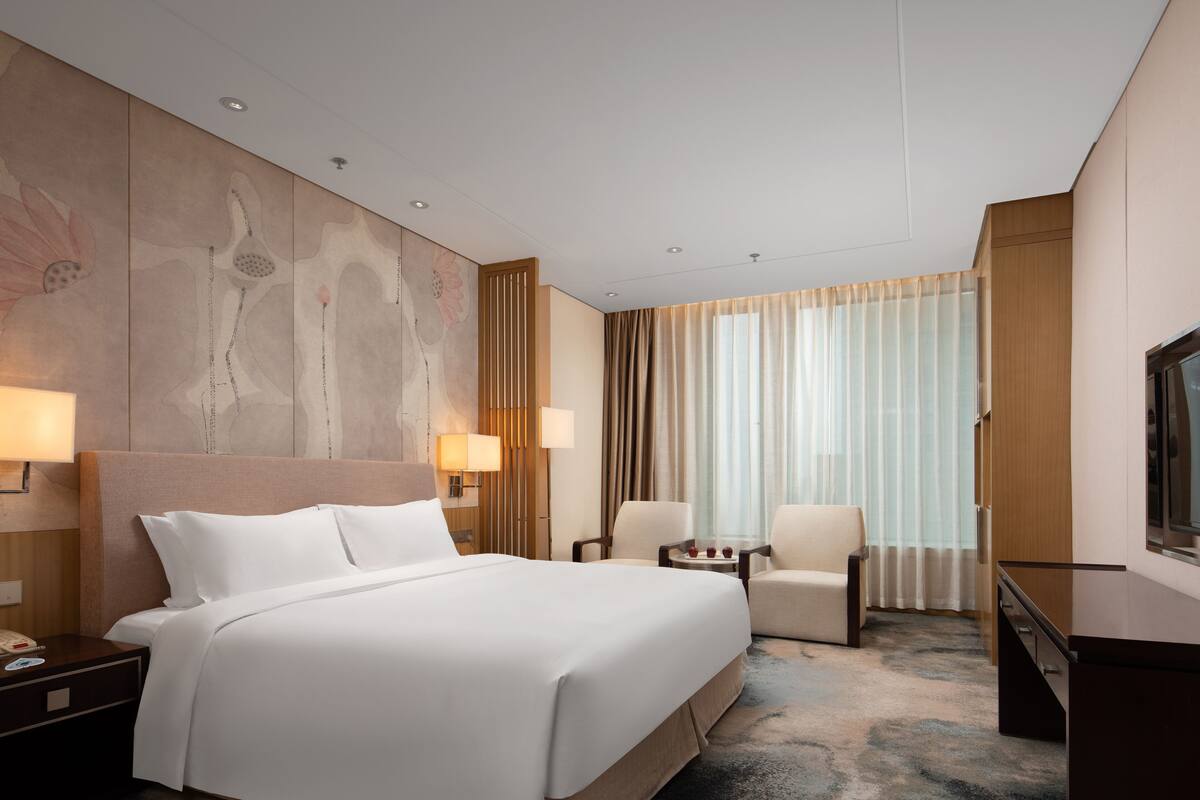 Quarto de hotel com uma cama grande, duas luminárias de cabeceira, uma escrivaninha com telefone, uma cadeira e uma televisão.