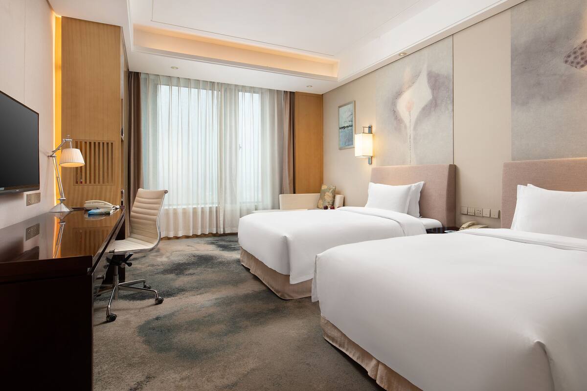 Quarto de hotel com duas camas, uma escrivaninha com luminária, uma cadeira, uma televisão e uma janela grande com cortinas.